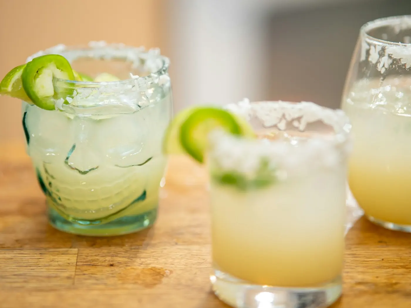 Two salt-rimmed margaritas with jalapeño slices on a wood counter