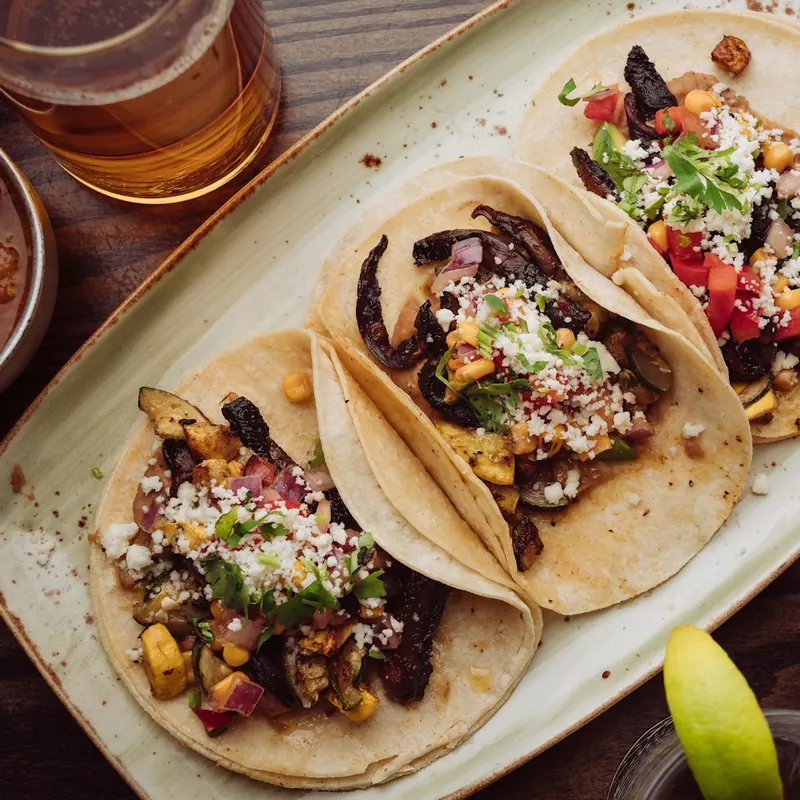 Three tacos with corn and cheese on a long plate beside a glass of beer