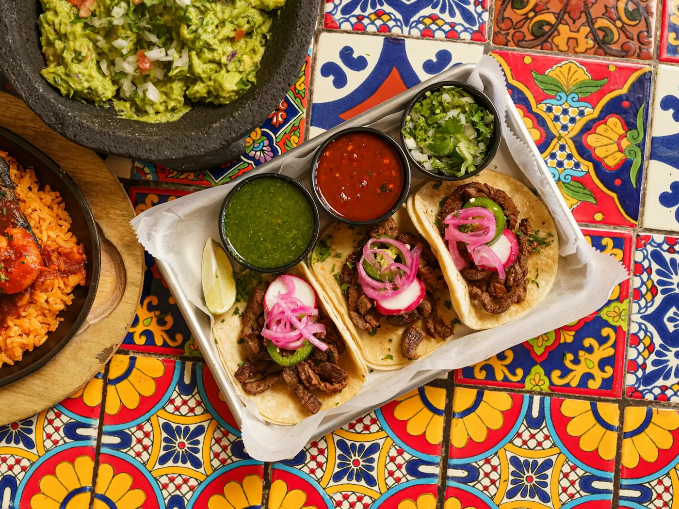 Three tacos with pickled red onion and four salsas on a tiled table